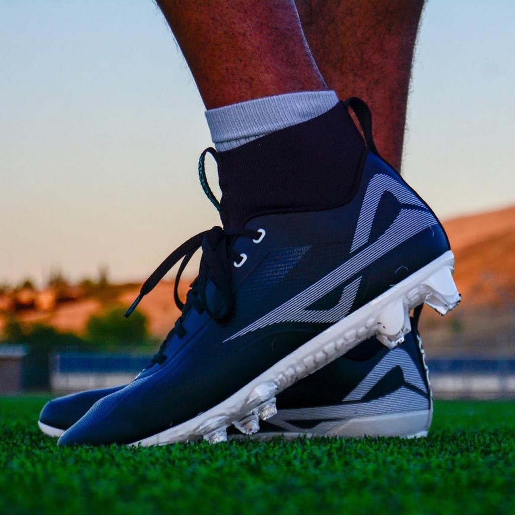 Athlete-on-turf photo of black/white Caddix mid men’s football & lacrosse cleats that use employ patented Smart Studs to reduce ankle injuries and ACL tears