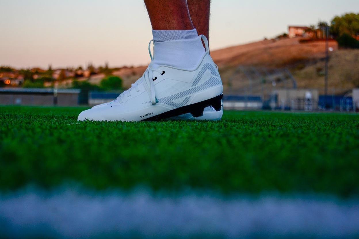 On field photo of athlete wearing Caddix men’s white/black mid football & lacrosse cleats, with patented Smart Studs that reduce ACL tears and ankle injuries
