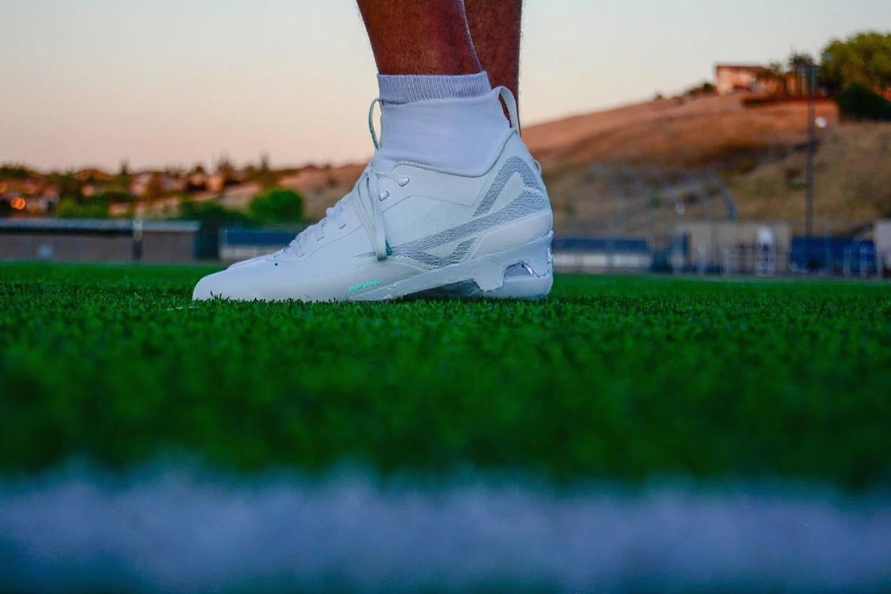 On the turf picture of white/white mid men’s Caddix football & lacrosse cleats that use patented Smart Studs to reduce ankle injuries and ACL tears